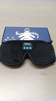 Front view of black YRITOS 3D sleep mask with Bluetooth control panel on top, placed on table with blue graphic box behind.