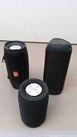 Three black portable Bluetooth speakers with mesh exterior. One branded MusiBaby on metallic top, one with orange G logo and fabric handle, and one trapezoidal shaped speaker with mesh grille.