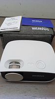 WiMius S25 white mini projector with lens and focus knob, user manuals and grey soft carrying case on a wooden surface.