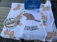White Canadian Centennial tablecloth with beaver and autumn leaves and the words 'Canadian Centennial' displayed on a couch with a commemorative Confederation plate.