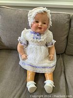 Full view of the vintage composition and cloth doll, dressed in white dress, bonnet, socks and shoes, positioned sitting on a couch.