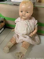Front view of the 22-inch composition and cloth baby doll seated, wearing a pink dress and cream knitted socks, showing signs of wear and paint loss on limbs.