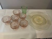 Photo shows six pink ribbed dessert bowls arranged in front, a yellow Depression glass platter with handles in the back center, and a tall green glass on the right side.