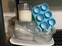 Tommee Tippee microwave baby bottle sterilizer container with multiple compartments, clear plastic colander, green Tupperware bowl, and various other kitchen items inside the clear trays.