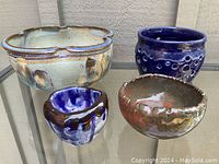 Four pottery bowls in various colors and sizes displayed on a glass surface, showing diverse glazing styles and textures.