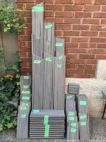 Composite decking boards of various lengths stacked vertically against brick wall with measurement tags.
