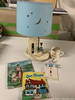 Lamp and items arranged on table, showing front view of lamp with blue lampshade decorated with stars and moon cutouts, two children's books and a cup.