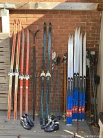 Six pairs of cross-country skis with matching poles and two pairs of ski boots placed against a brick wall; skis include vintage waxable and modern waxless styles.