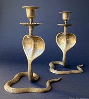Front view of two brass cobra-shaped candle holders showing coiled snake bases and raised engraved hoods