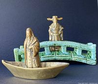 Brass figurines, brass boat, and ceramic bridge arranged against black backdrop