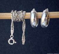 Spiral chain bracelet coiled next to pair of silver hoop earrings on a wooden rod, all set against a dark background showing to-scale size and clasp detail of bracelet and shape of earrings.