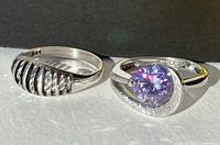 Two silver rings, one with a large lavender cubic zirconia and one with an openwork design, shown on flat surface with sunlight highlighting.