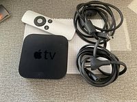 Apple TV 3rd generation device, white remote control, black power cable, and black HDMI cable arranged on a carpeted surface.