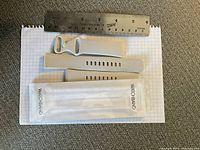 Two beige Fitbit watchbands displayed on graph paper with a metal ruler showing their length and a plastic sleeve packaging beside them.