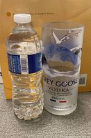 Clear cylindrical glass with Grey Goose Vodka branding and mountain and geese design next to a water bottle for scale.