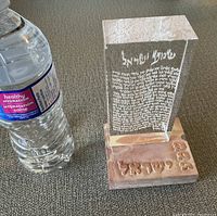Clear acrylic Judaica stand next to water bottle for size reference; acrylic block engraved with Hebrew prayer text on a stone base with Hebrew lettering.