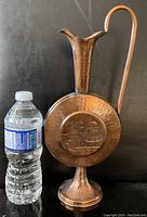 Copper jug with hammered texture and round embossed Jerusalem design, standing next to a water bottle for scale.