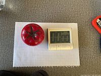 Top-down photo of two kitchen alarm timers on a grid paper sheet: a red tomato-shaped mechanical timer and a white digital timer with buttons.