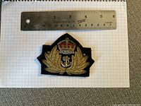 Embroidered Royal Navy officer cap badge placed on graph paper with a ruler above it, showing its size approx 4 cm wide.