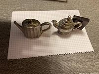 Two vintage teapot-shaped salt and pepper holders on graph paper; one ribbed metal style, one silver-toned with engraved design
