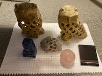 Overview of five stone figurines including two hollow carved owls, a blue carved animal, a grayish carved figurine, and a rose quartz sphere next to matchbox for scale