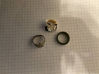 Photo showing three rings on a grid paper background: one silver marked ring with a subtle design, one green jade ring, and one black and white shell ring with a marbled pattern.