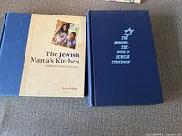 Two hardcover Jewish cookbooks side by side on a grey carpet: 'The Jewish Mama's Kitchen' with a beige and blue cover and 'The Around-The-World Jewish Cookbook' with a dark blue cover and star design.