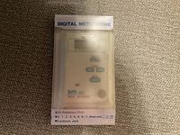 Digital metronome in original clear plastic box on carpeted surface showing brand, model, and control buttons.