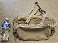 Front view of beige canvas bag with zippered pocket and handles, alongside a water bottle for size reference.