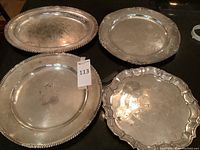 Four silver plate serving platters placed on black surface, showing varied designs and heavy surface wear.