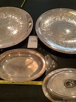 Four silver plate serving pieces on black surface: two large engraved trays, one small oval tray, and a Gorham covered serving dish with handle.