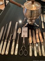 Full view of the silver plate fondue pot on its tripod stand with attached fuel burner, alongside a collection of mother of pearl utensils including knives, forks, a spoon, and ornate metal scissors.