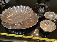 Overview of silver plated lot showing two large scalloped edge trays stacked, two small bowls, and a trophy form cup with two handles.