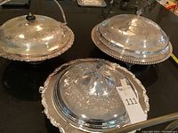 Photo showing all three silver plated lidded serving dishes arranged together on a black surface, showing decorative engravings and handles.