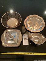 All four silver plated serving pieces displayed on a black surface: wire bread basket, scalloped round dish, square covered dish with lid, and gravy boat.