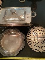 Silver plated chafing dish with hinged lid, round serving tray, and expandable trivet placed together on a black surface.