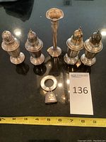 Full overview of all silver items on black surface including four salt and pepper shakers, bud vase, bell, measuring tape.