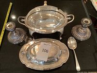 Photo showing two silver weighted candlesticks, silver serving dish with handles and hinged lid, silver covered serving dish on tray, and sterling silver serving spoon on black surface with measuring tape.