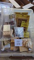 Clear plastic bin containing assorted dollhouse wooden components including packaged windows, trim, spindles, numbered with lot tag.