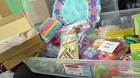 Photo of a plastic bin containing various party items including floral napkin rings, colorful stir sticks, seashells, candles, and a scallop-edge plate with a tropical pattern.