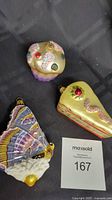 Three blown glass ornaments including cupcake, cake slice, and butterfly on a black surface with lot number card