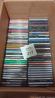 Box containing approximately 50 music CDs in jewel cases, spines visible showing assorted artist names and genres