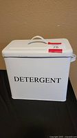 Front view of white rectangular metal detergent tin with 'DETERGENT' text and top handle.