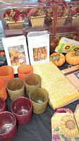 Fall themed decor items arranged including votive holders in orange, green, red, and gold, floating candles packaged, yellow patterned table runner, faux pumpkins, sunflower napkins, and gift tags