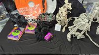 Photo showing a group of Halloween decorations: three white articulating skeletons in seated and standing poses, spider web themed plates, black rat skeletons, black raven figurines, and parts of orange paper lanterns in the background.