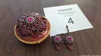 Round enameled box with magnetized brooch and dangling earrings displayed on wooden surface