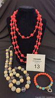 Full view of three jewelry pieces on black bust and display including two necklaces (a long coral-colored multi-strand bead necklace and a faceted bead crystal-accents necklace) and one beaded bracelet with turquoise and gold accents.