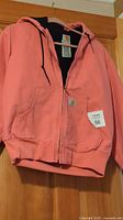 Full front view of salmon colored ladies Carhartt jacket hanging on hanger. Shows zip closure, hood with drawstrings, kangaroo pockets and ribbed cuffs and waistband.