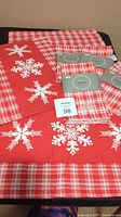 Red fabric placemats with white snowflakes and red plaid edging alongside packaged red plaid cloth napkins embroidered with white snowflakes.