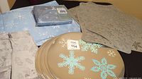 Photo showing light blue snowflake tablecloth in packaging, beige/taupe cloth placemats and napkins, and taupe rectangular plastic placemats with cutout snowflake designs.